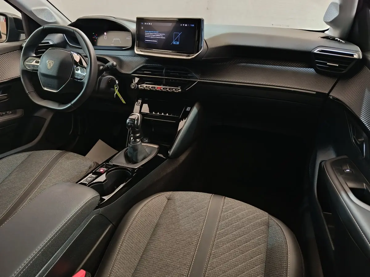 Intérieur avant de la Peugeot 208 PureTech 100 S&S Allure 2025 gris foncé, avec volant cuir et écran tactile central.