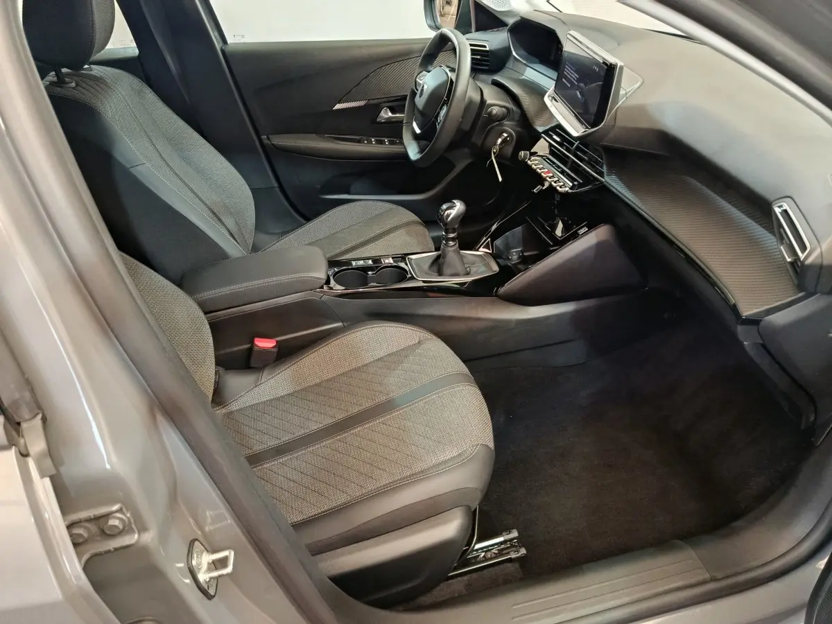 Vue intérieure côté conducteur de la Peugeot 208 PureTech 100 S&S Allure 2025 avec sièges gris et écran tactile central.