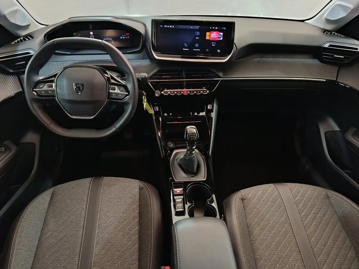 Vue intérieure avant de la Peugeot 208 PureTech 100 S&S Allure 2025, sièges gris et tableau de bord moderne avec écran tactile.