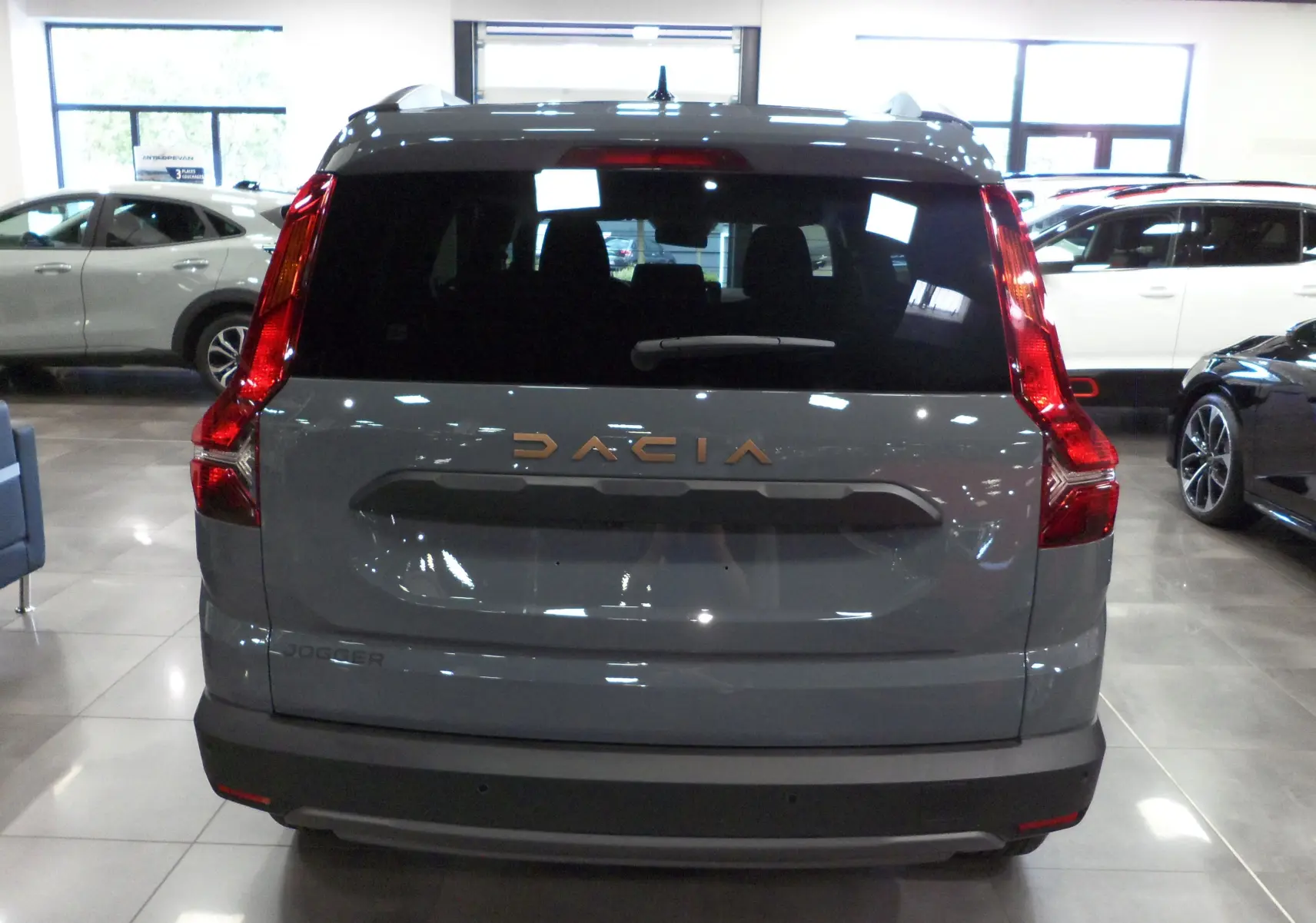 Vue arrière d'un Dacia Jogger gris urbain 2025 avec feux verticaux et logo doré dans un showroom.