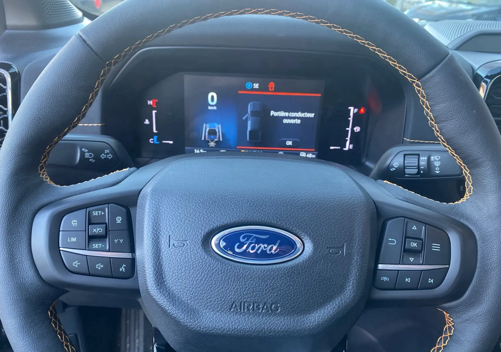Vue rapprochée du volant cuir noir avec surpiqûres orange du Ford Ranger 2025, tableau de bord numérique affichant une porte ouverte.