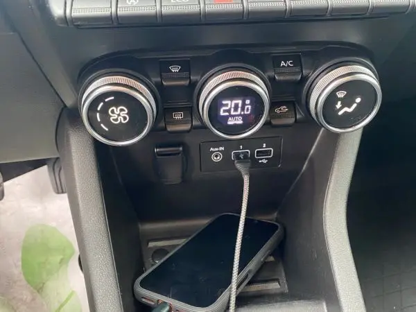 Gros plan sur la console centrale de la Renault Clio V gris platine, montrant les commandes de climatisation et un smartphone connecté.