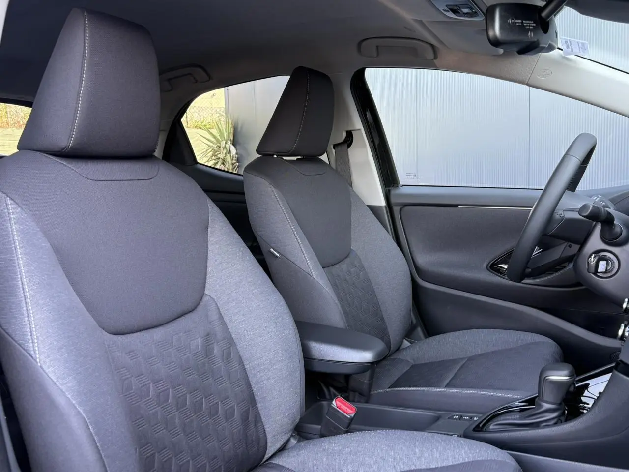 Vue intérieure côté conducteur de la Toyota Yaris 2025 hybride, sièges gris avec accoudoir central et volant cuir.