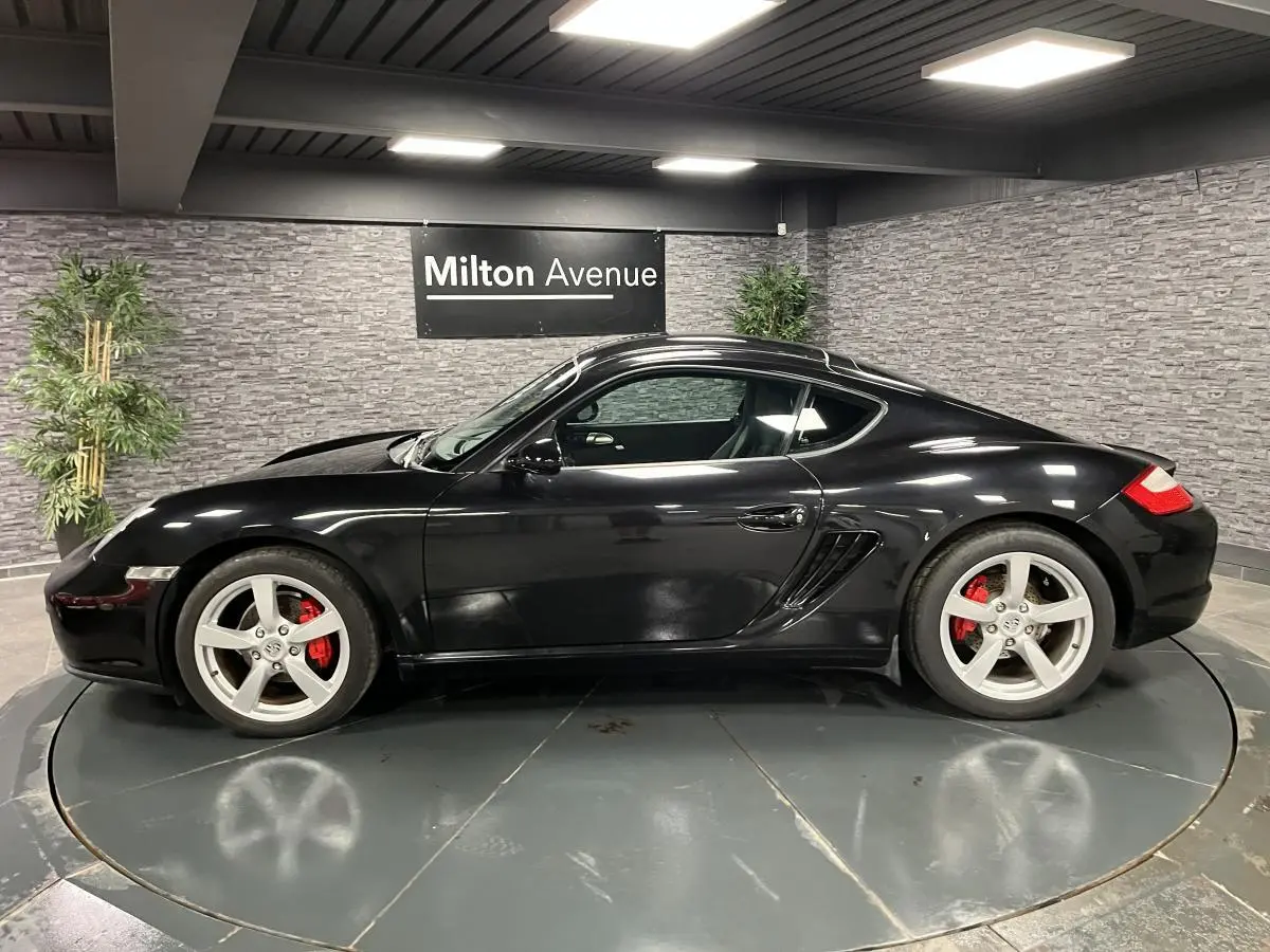Profil côté gauche d'une Porsche Cayman noire 2008 avec jantes argentées et étriers de frein rouges en intérieur showroom.