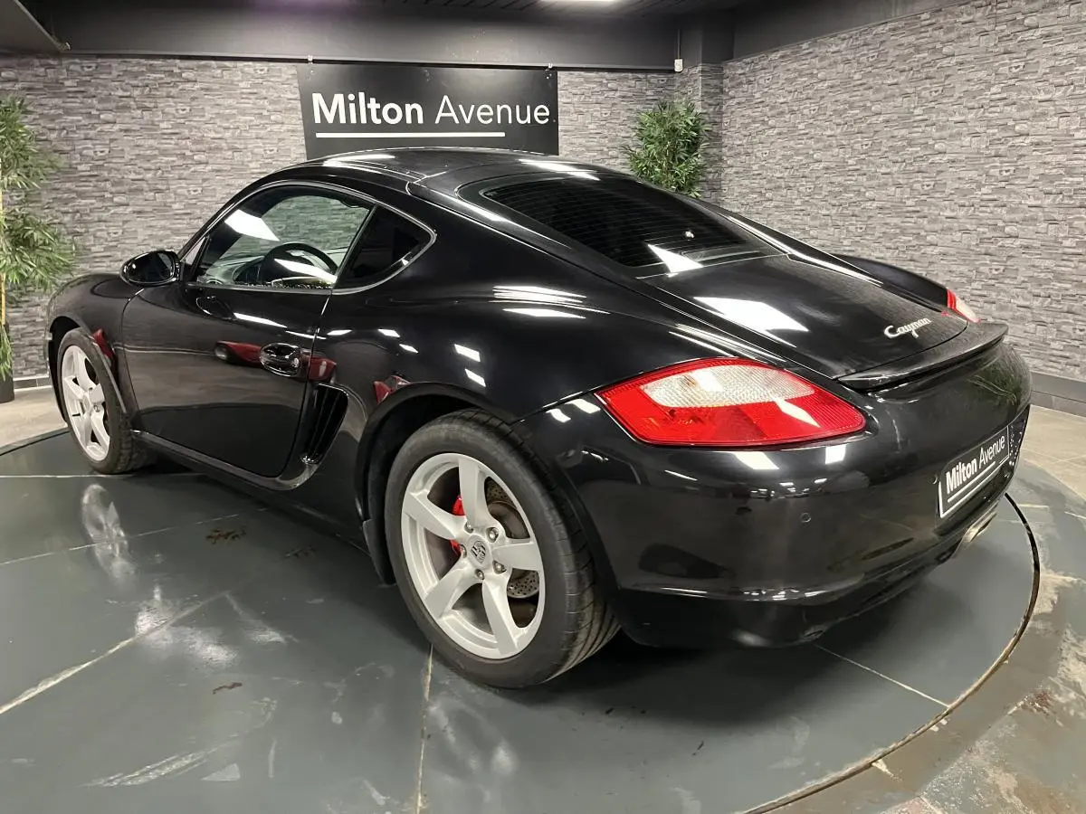 Vue 3/4 arrière droite d'une Porsche Cayman noire avec jantes argentées et feux arrière rouges, en intérieur showroom.
