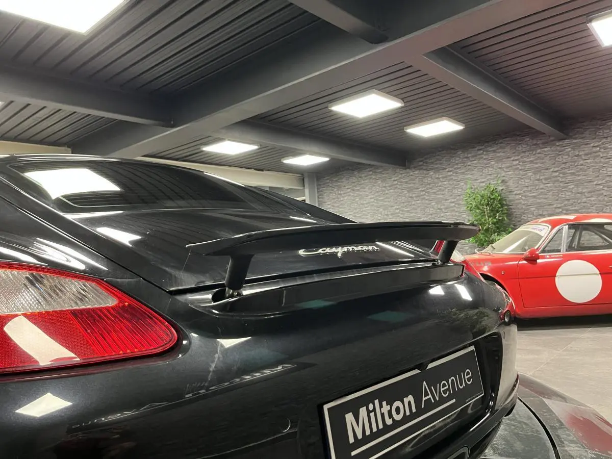 Vue arrière droite d'une Porsche Cayman noire avec aileron arrière déployé dans un showroom intérieur.