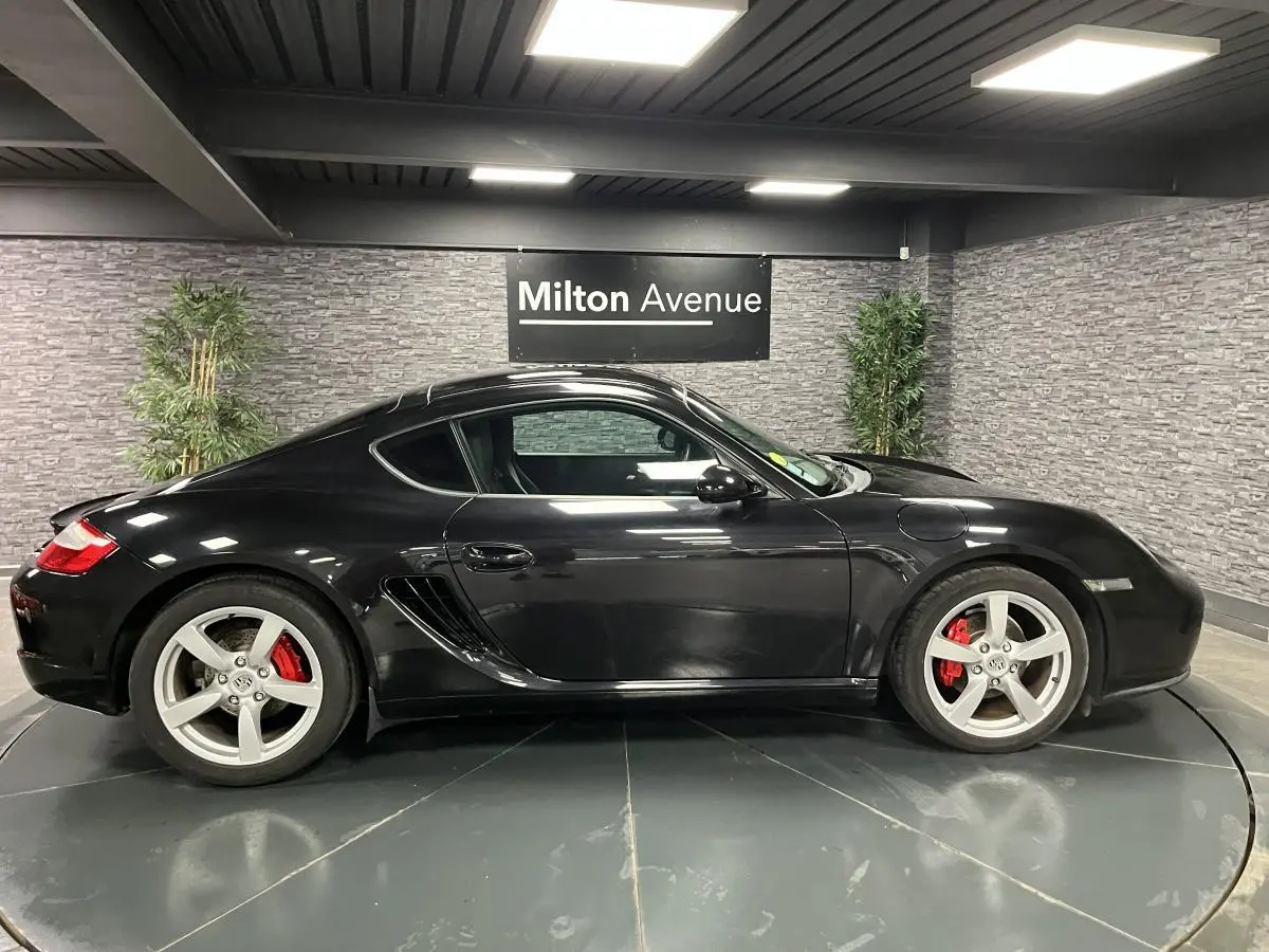 Profil côté gauche d'une Porsche Cayman noire 2008 avec jantes argentées et étriers de frein rouges en intérieur showroom.