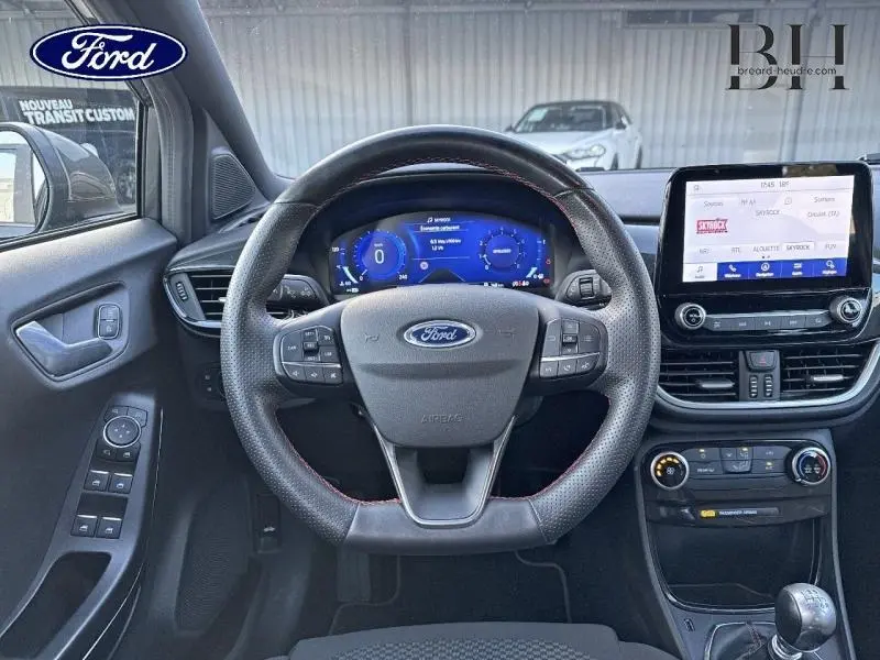 Vue intérieure centrée sur le volant sport et le tableau de bord numérique du Ford Puma gris avec écran tactile et commandes au volant.
