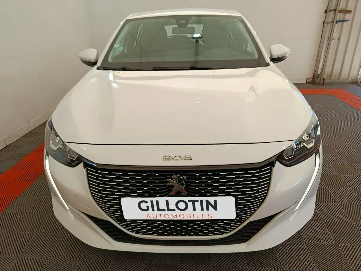 Vue avant d'une Peugeot 208 électrique blanche avec calandre noire et feux LED, en intérieur sur sol à damier rouge et gris.