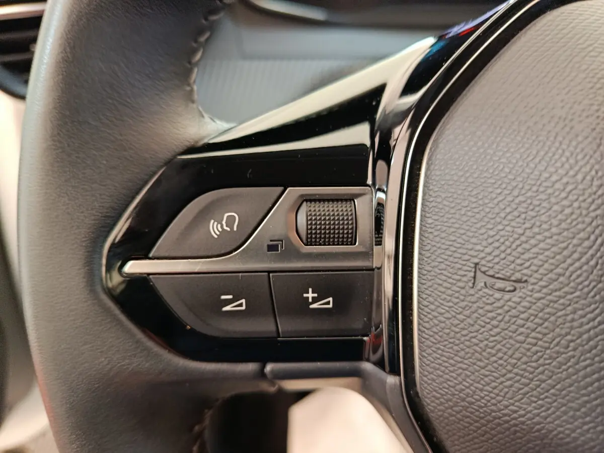 Gros plan sur les commandes gauche du volant cuir noir du Peugeot 208 électrique blanc, avec boutons multifonctions.