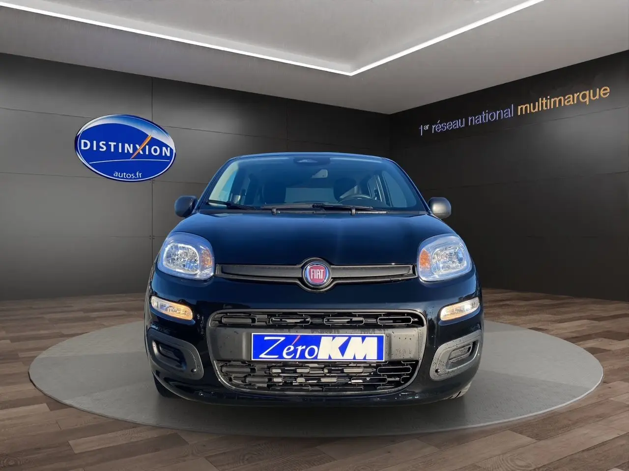 Vue frontale d'une FIAT Panda noire 2025 avec phares allumés et plaque "Zéro KM" en showroom.