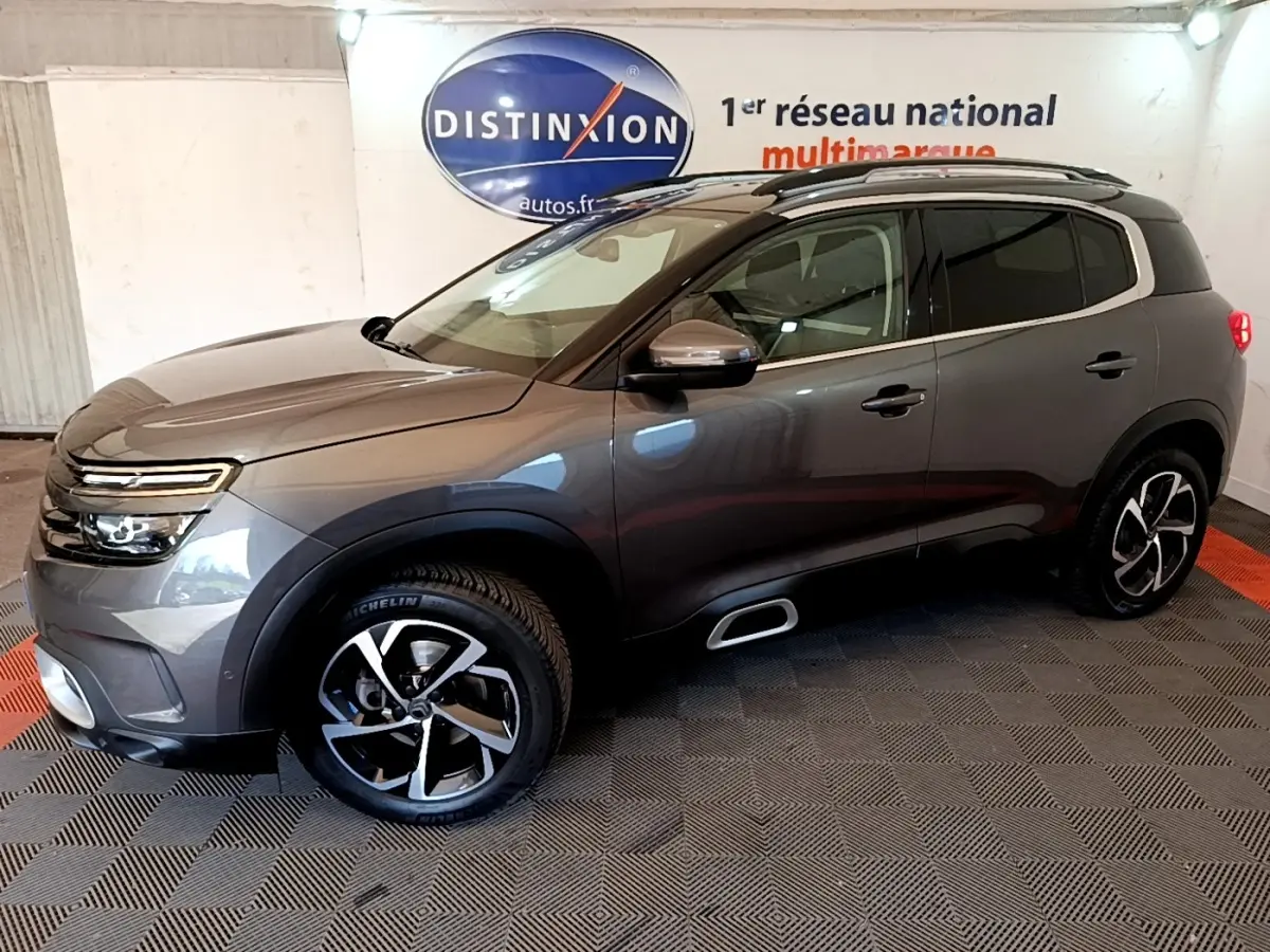 Vue de profil droit du Citroën C5 Aircross gris foncé avec jantes alliage et barres de toit chromées en intérieur showroom.