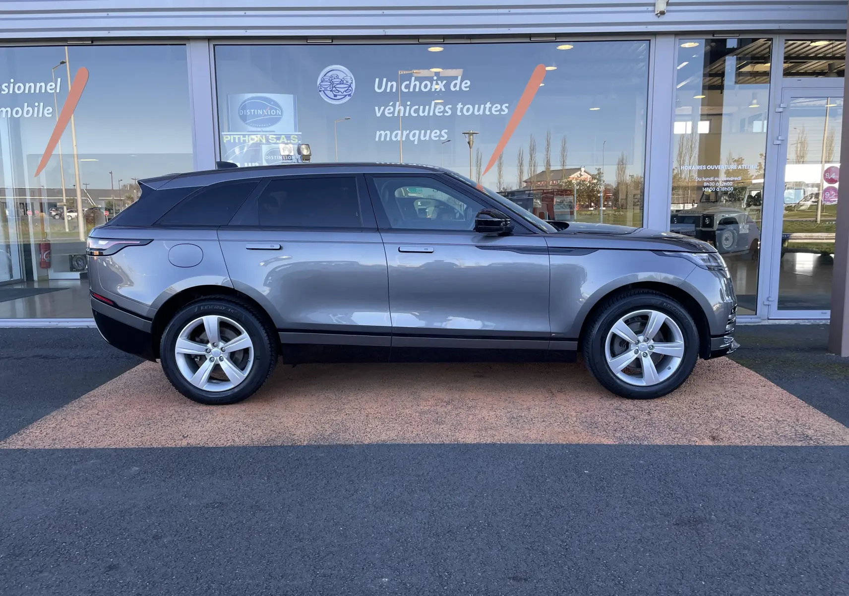Profil côté gauche d'un Range Rover Velar D240 S R-Dynamic gris Corris, avec jantes alliage et toit noir devant un showroom.
