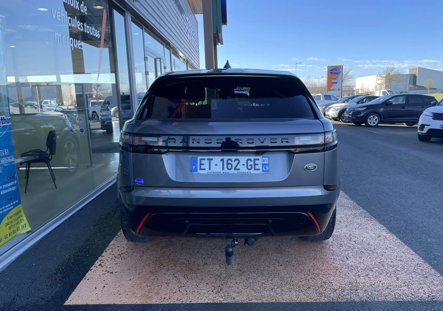 Vue arrière d'un Range Rover Velar gris Corris avec attelage, stationné devant une vitrine de concession.