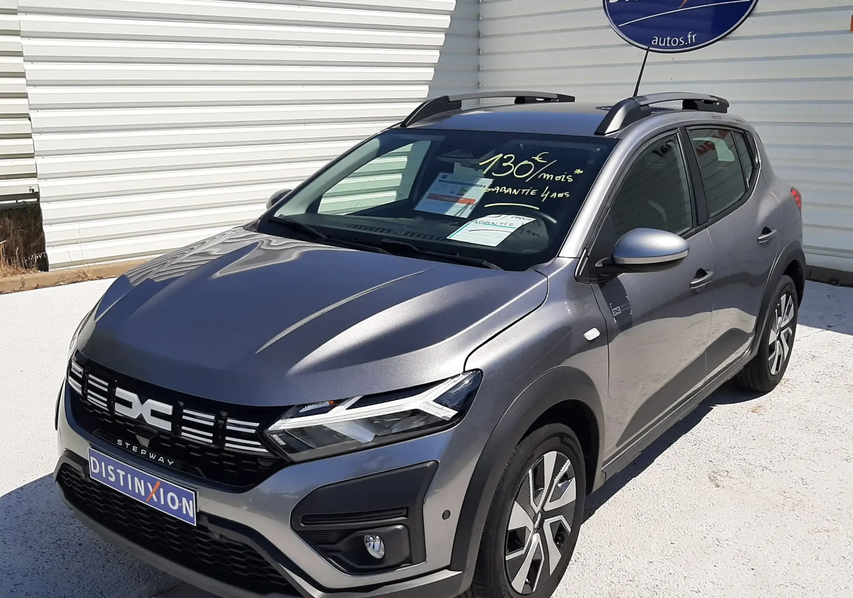 Dacia Sandero Stepway gris schiste vue 3/4 avant droit, avec barres de toit et stickers publicitaires visibles.