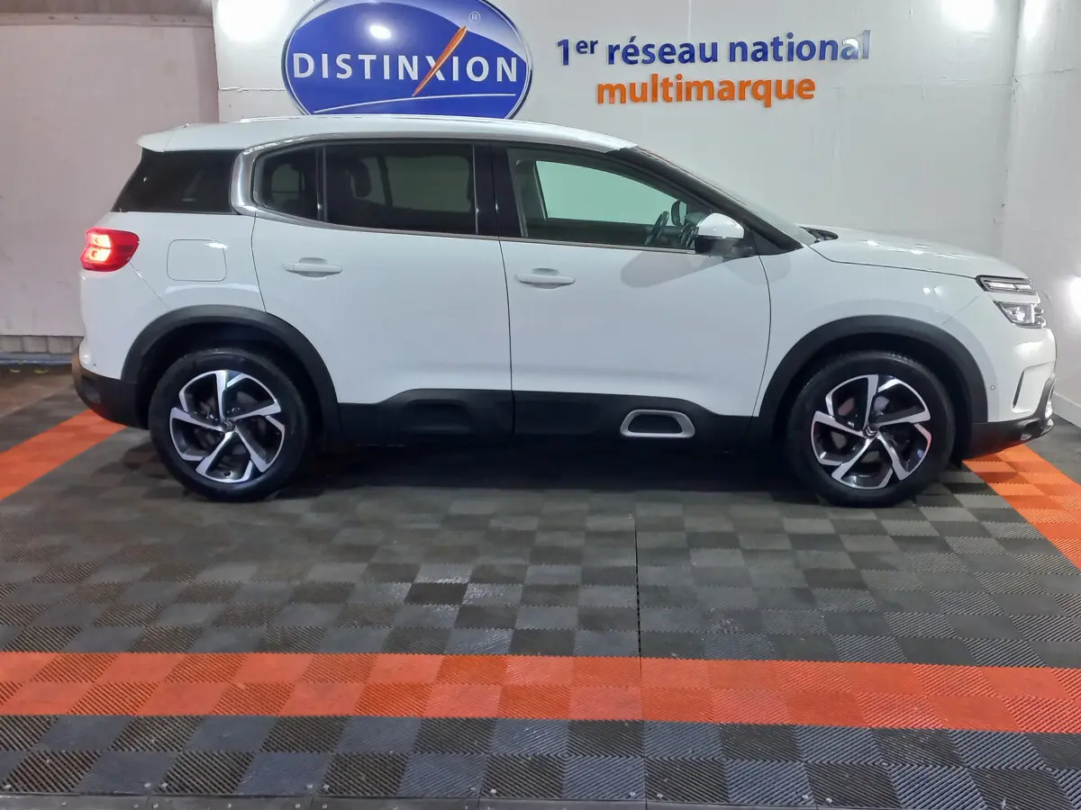 Profil droit d'un SUV Citroën C5 Aircross blanc, avec jantes alliage bicolores et feux arrière allumés en intérieur showroom.