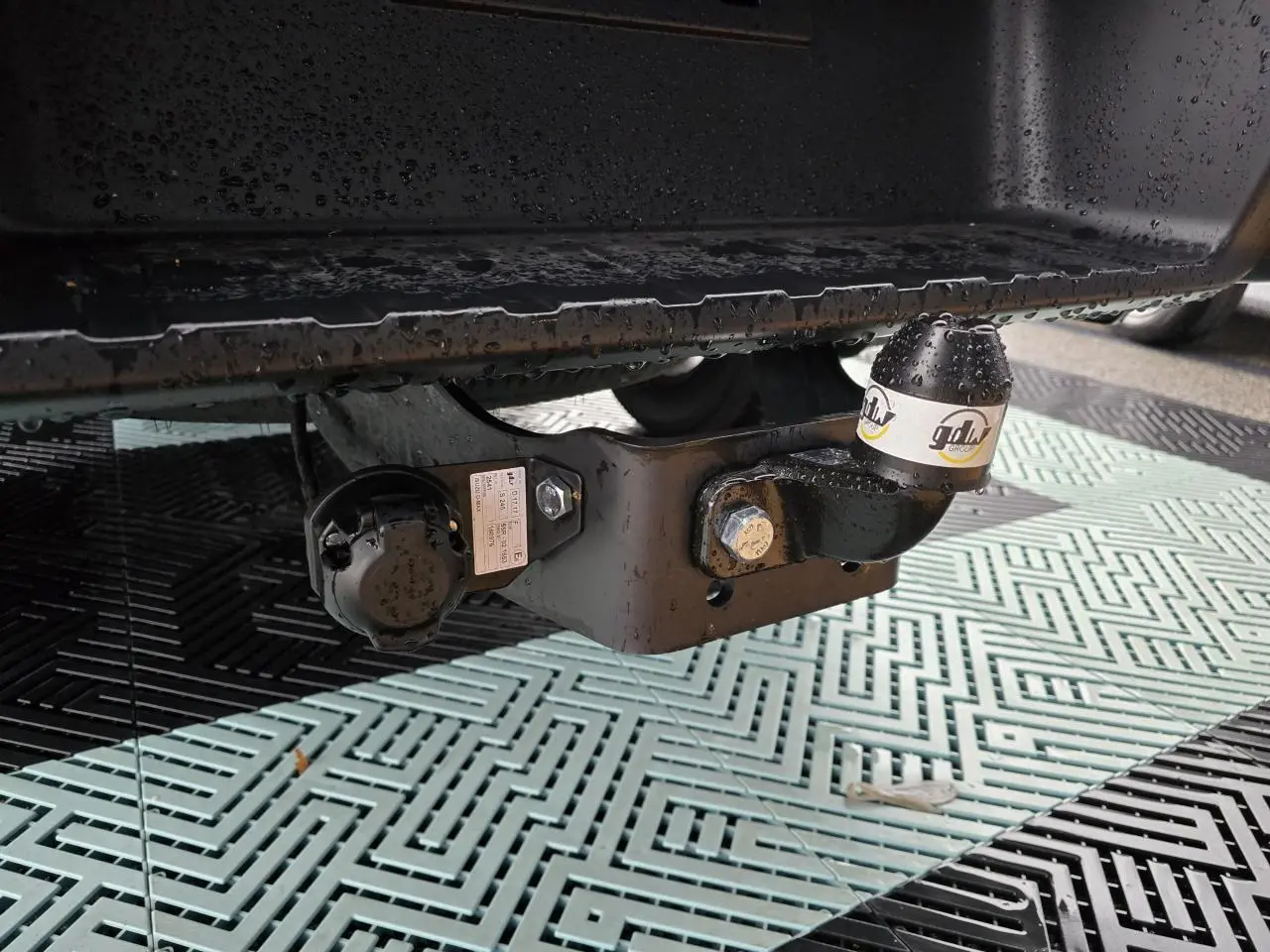 Gros plan sur l'attelage noir sous la benne d'un Isuzu D-MAX orange métallisé, avec bouchon de protection marqué gdlw.