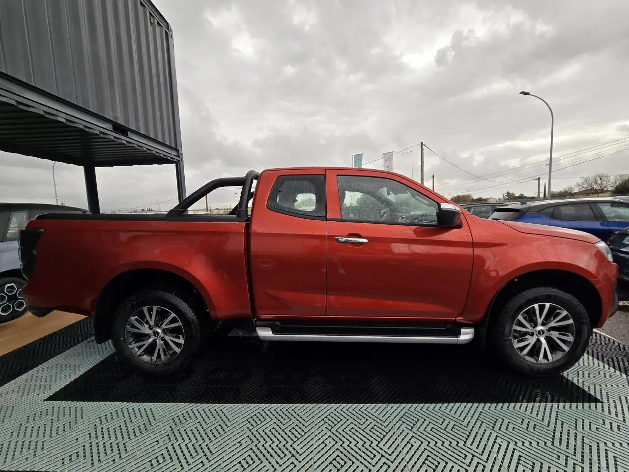 Vue de profil côté gauche d'un pick-up Isuzu D-MAX orange métallisé avec marchepieds argentés et arceau noir.