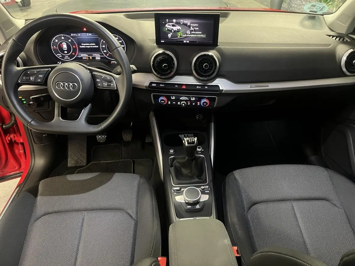 Intérieur Audi Q2 2024 rouge, vue de face sur tableau de bord, volant sport cuir, écran central et boîte manuelle.