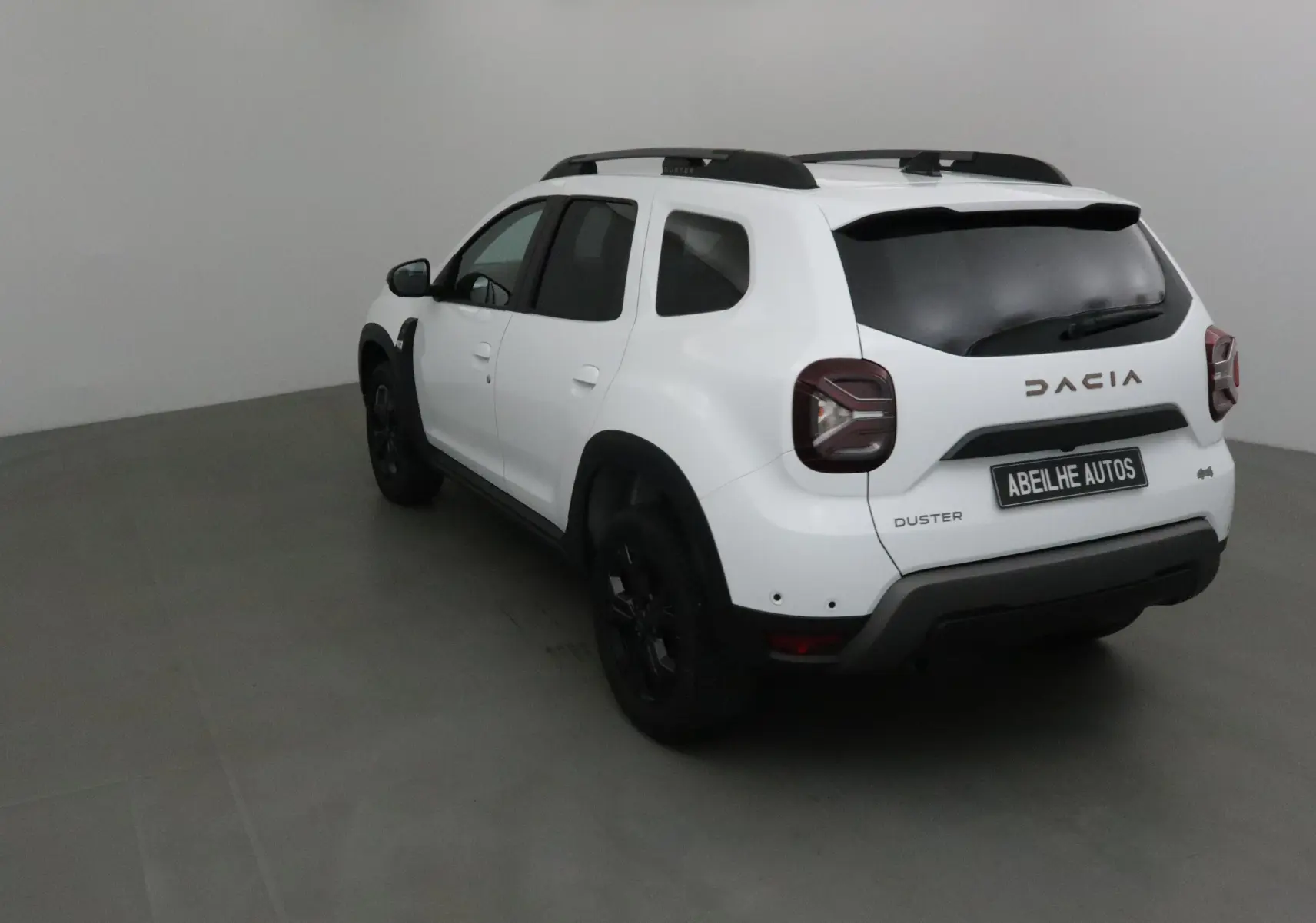 Vue 3/4 arrière droite d'un Dacia Duster blanc 2024 avec jantes noires et barres de toit noires.