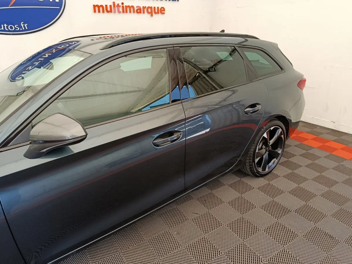 Vue latérale côté gauche d'une CUPRA LEON ST gris foncé avec jantes noires et toit ouvrant visible en intérieur showroom.