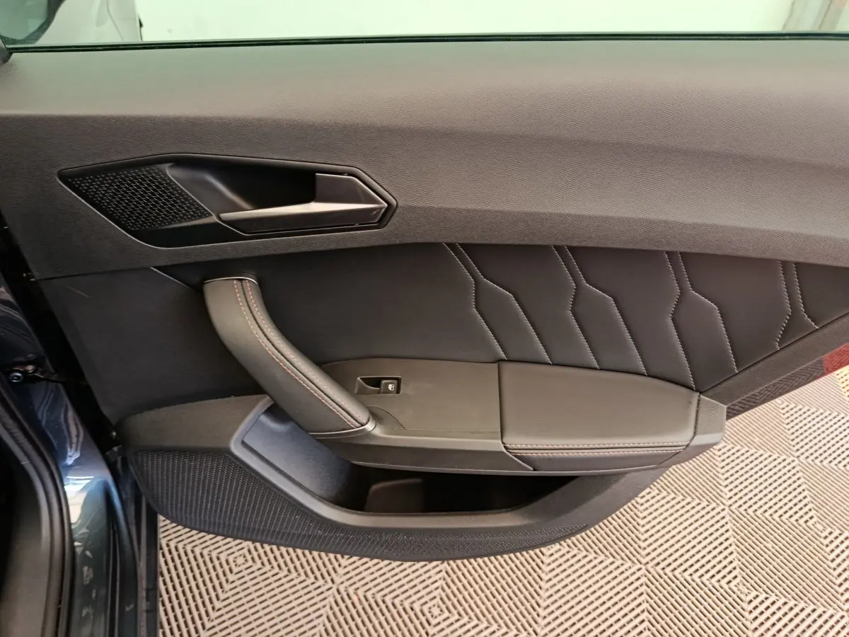 Vue intérieure côté gauche de la porte avant gris foncé de la CUPRA LEON ST avec poignée et surpiqûres rouges.