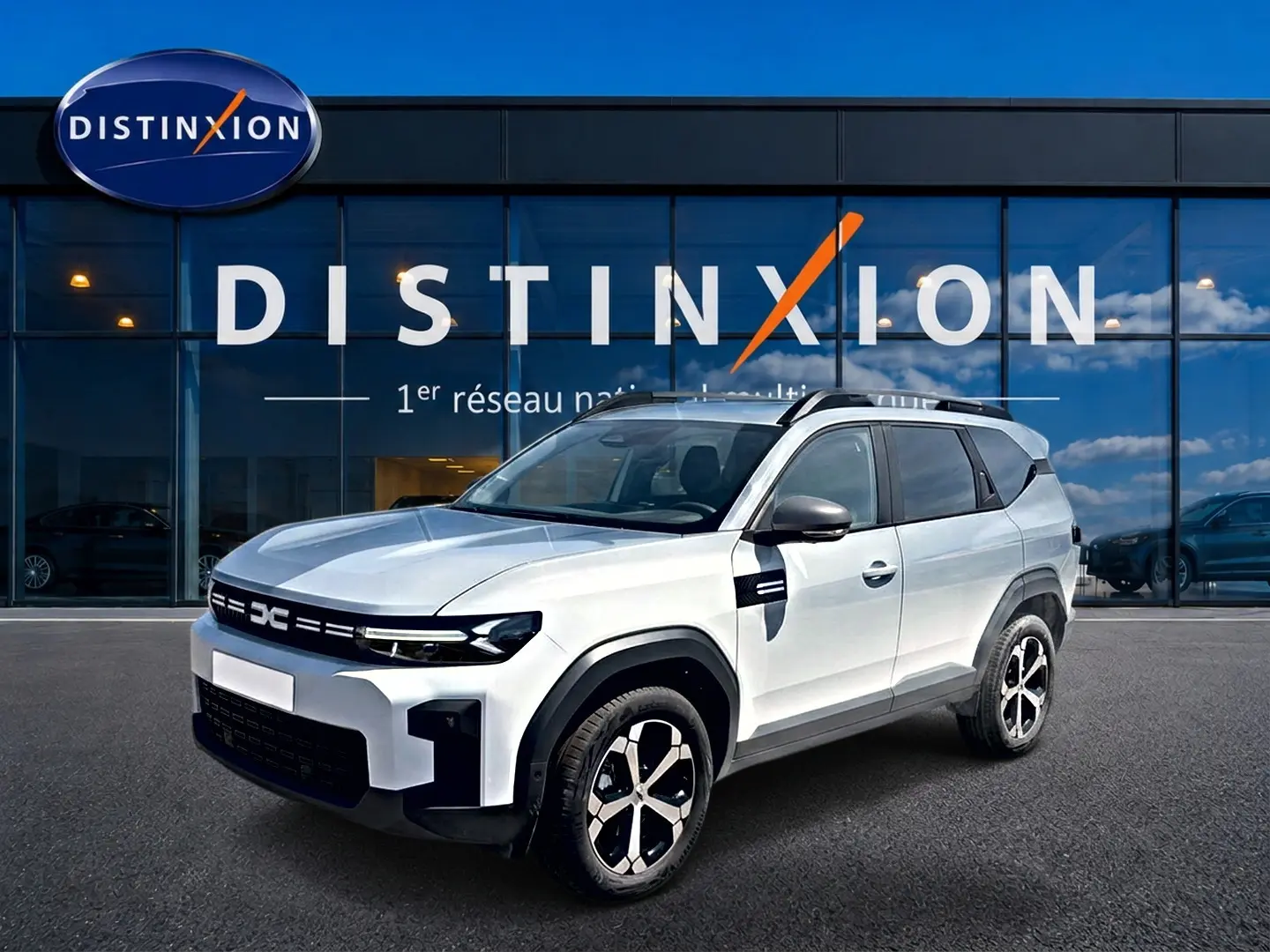 Vue 3/4 avant droit d'un Dacia Bigster blanc avec barres de toit et jantes alliage devant un bâtiment vitré Distinxion.