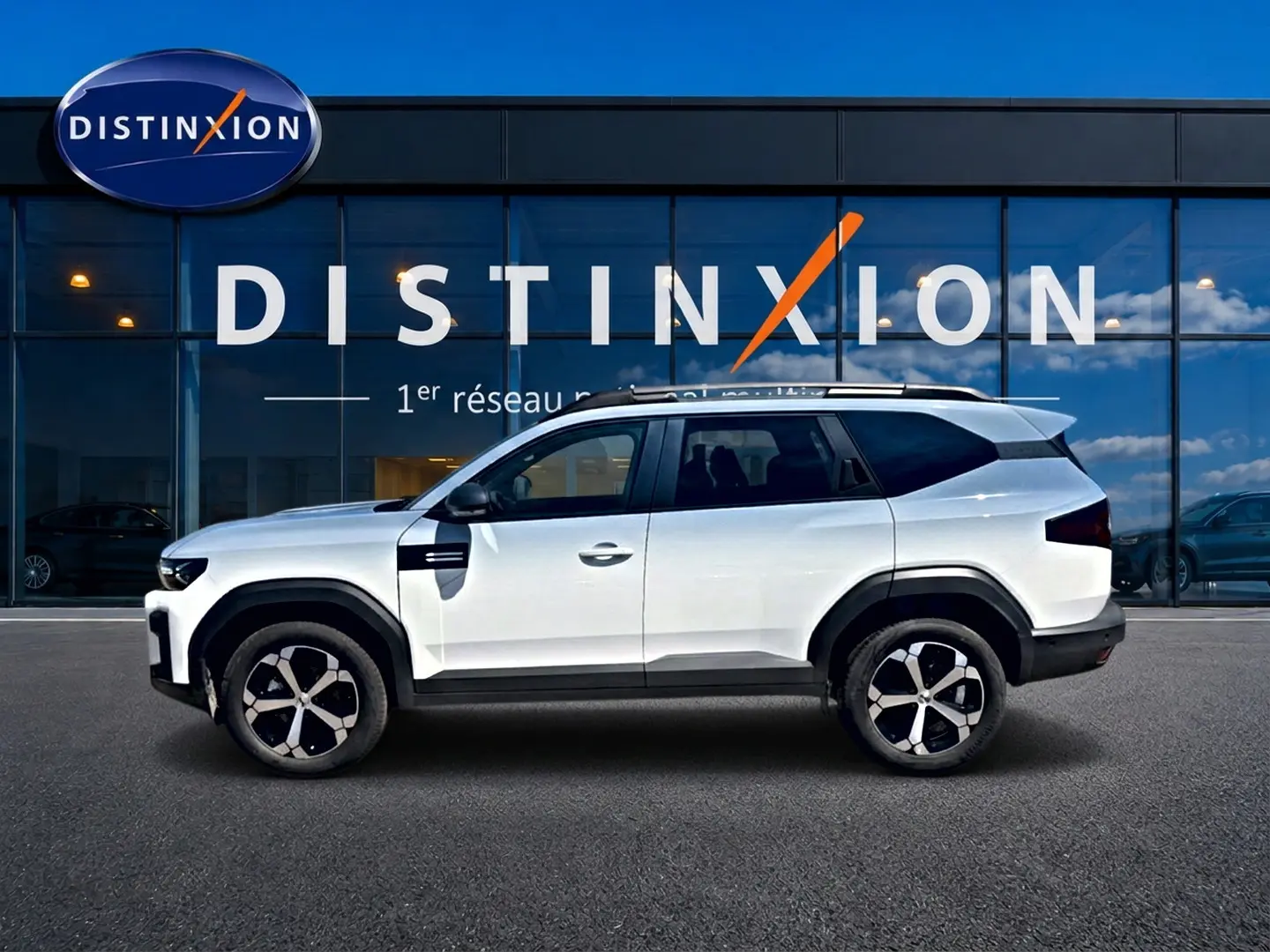 Profil droit du Dacia Bigster blanc 2026 avec jantes alu et barres de toit devant une vitrine Distinxion.