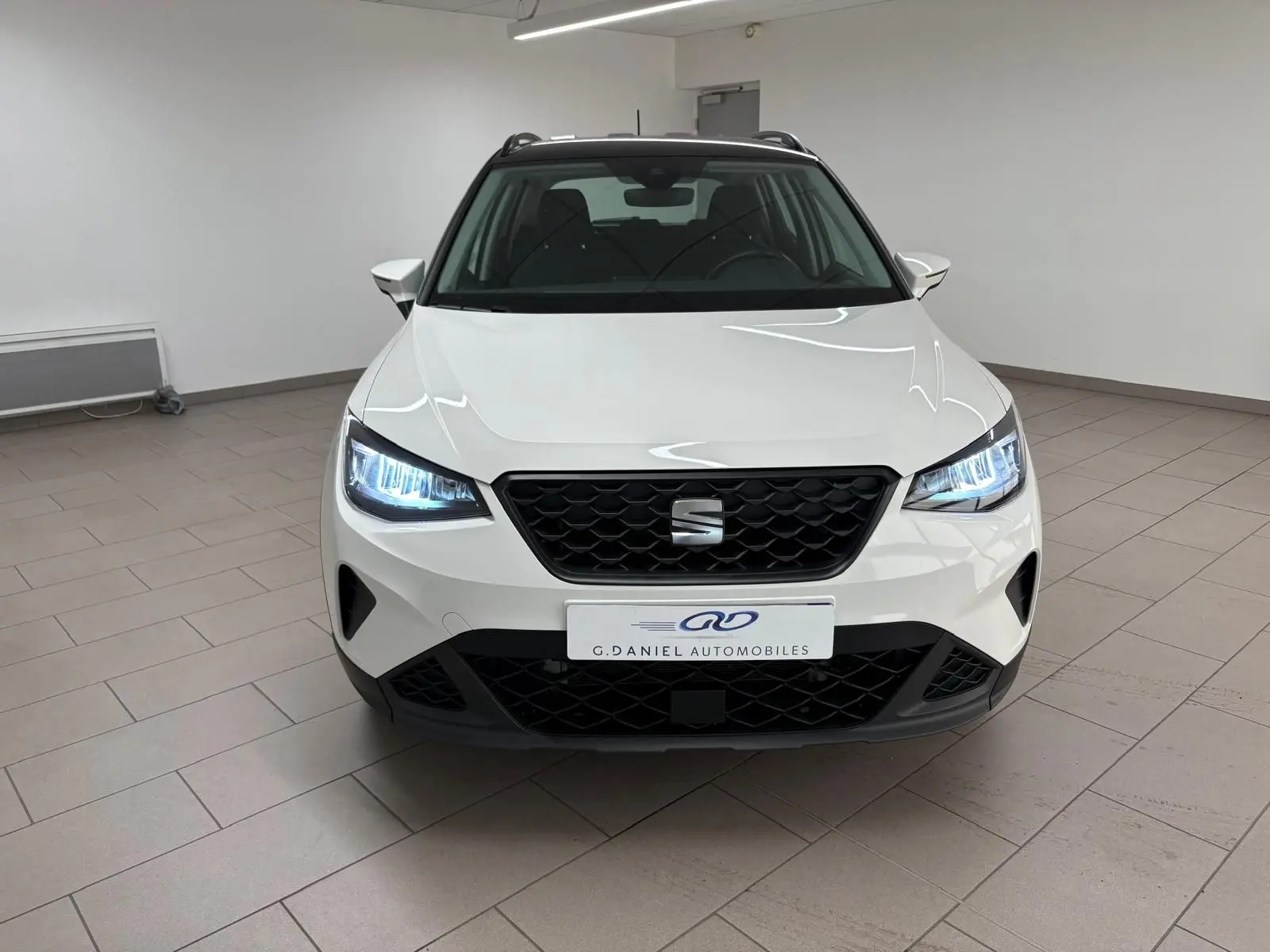 Vue de face d'une SEAT Arona 2023 blanche avec toit noir, phares allumés dans un intérieur neutre.