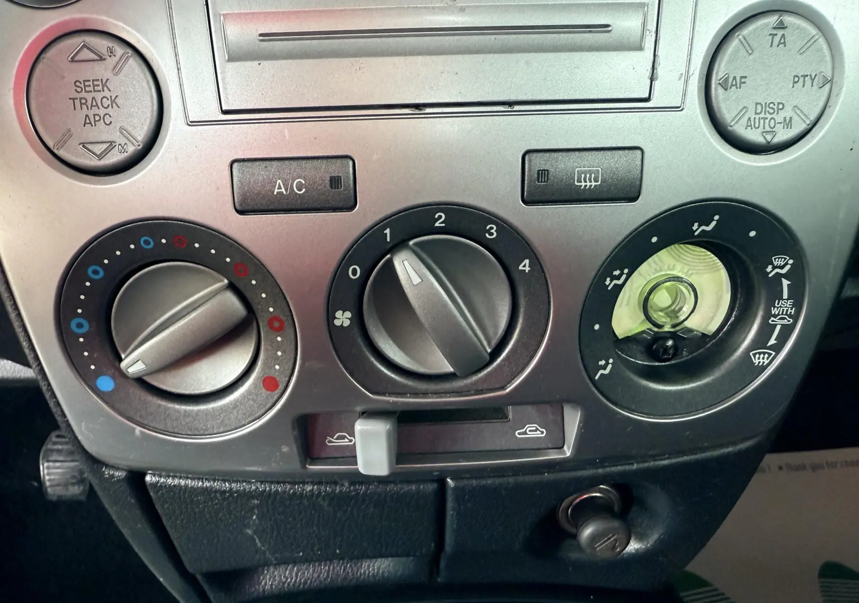 Gros plan sur les commandes de climatisation et radio argentées de la Mazda2 gris 2004, intérieur noir.