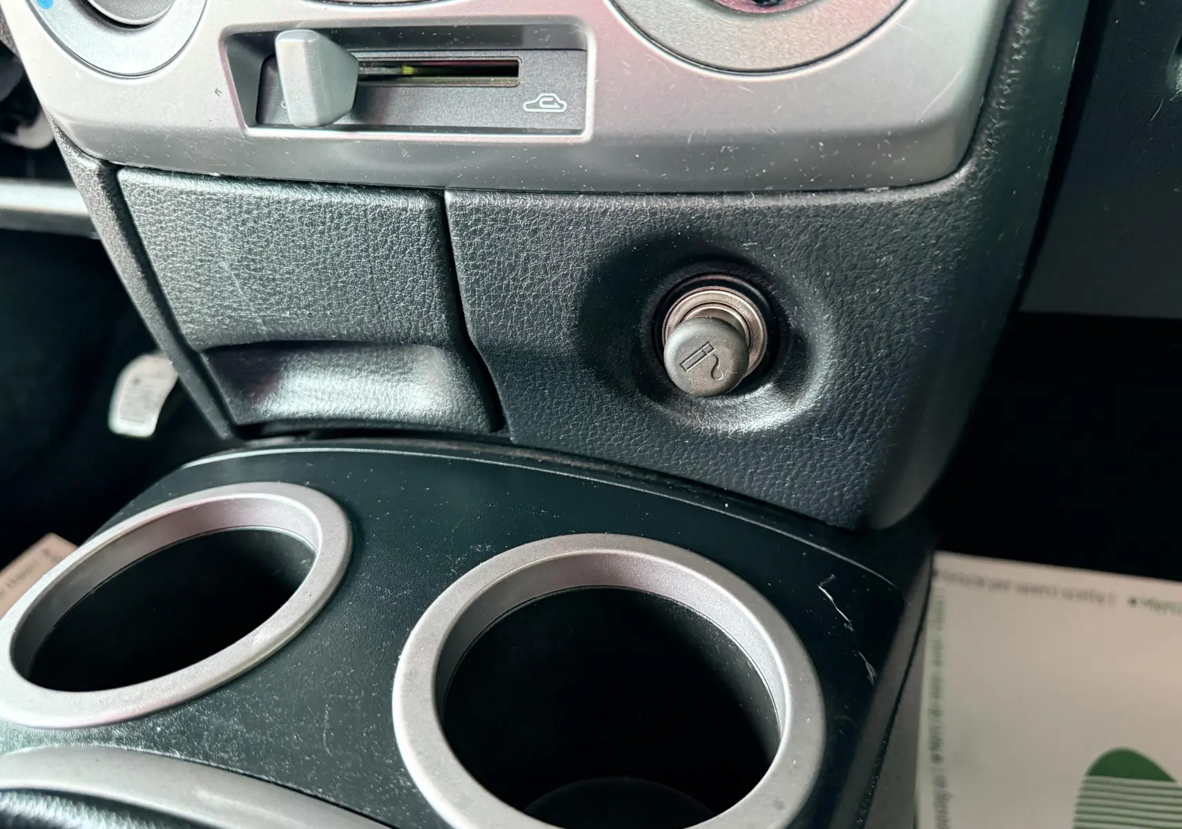 Gros plan sur la console centrale noire de la Mazda2 2004 montrant l'allume-cigare et les porte-gobelets argentés.