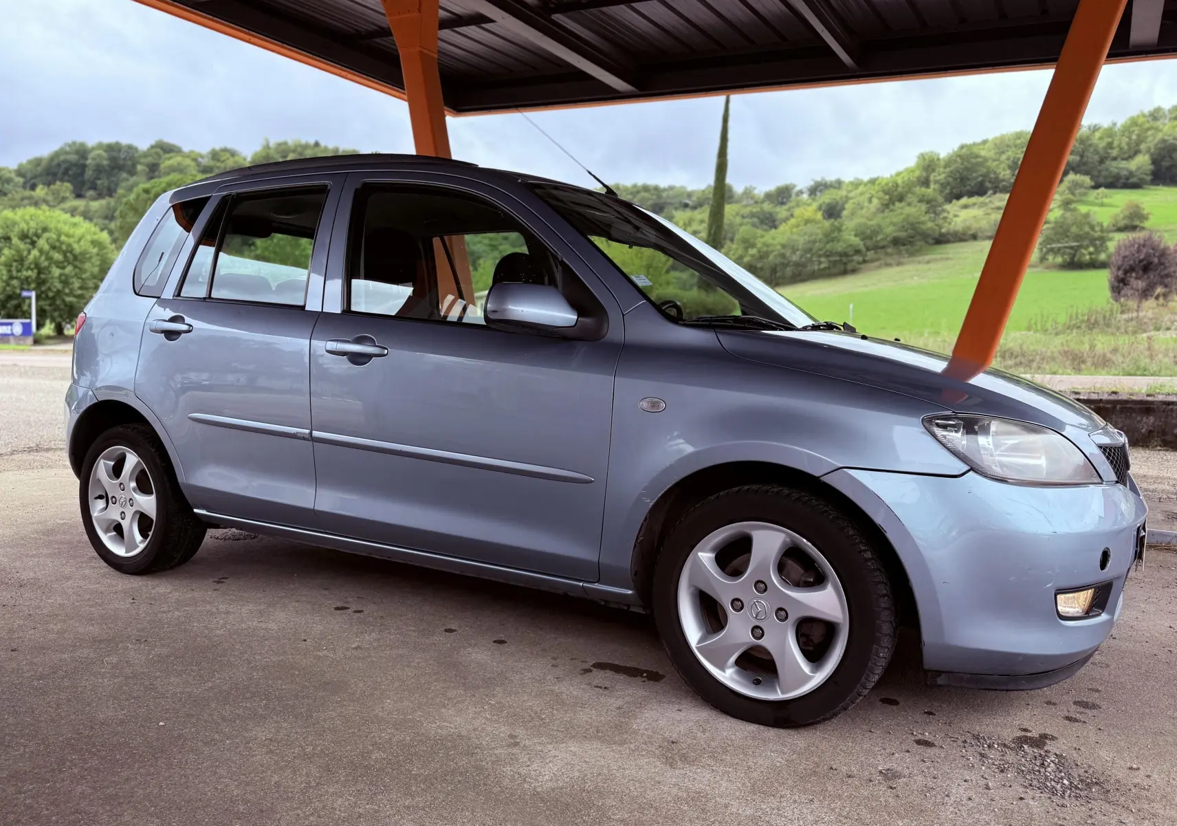 Vue de profil côté gauche d'une Mazda2 gris clair 5 portes garée sous un abri avec un paysage verdoyant en arrière-plan.