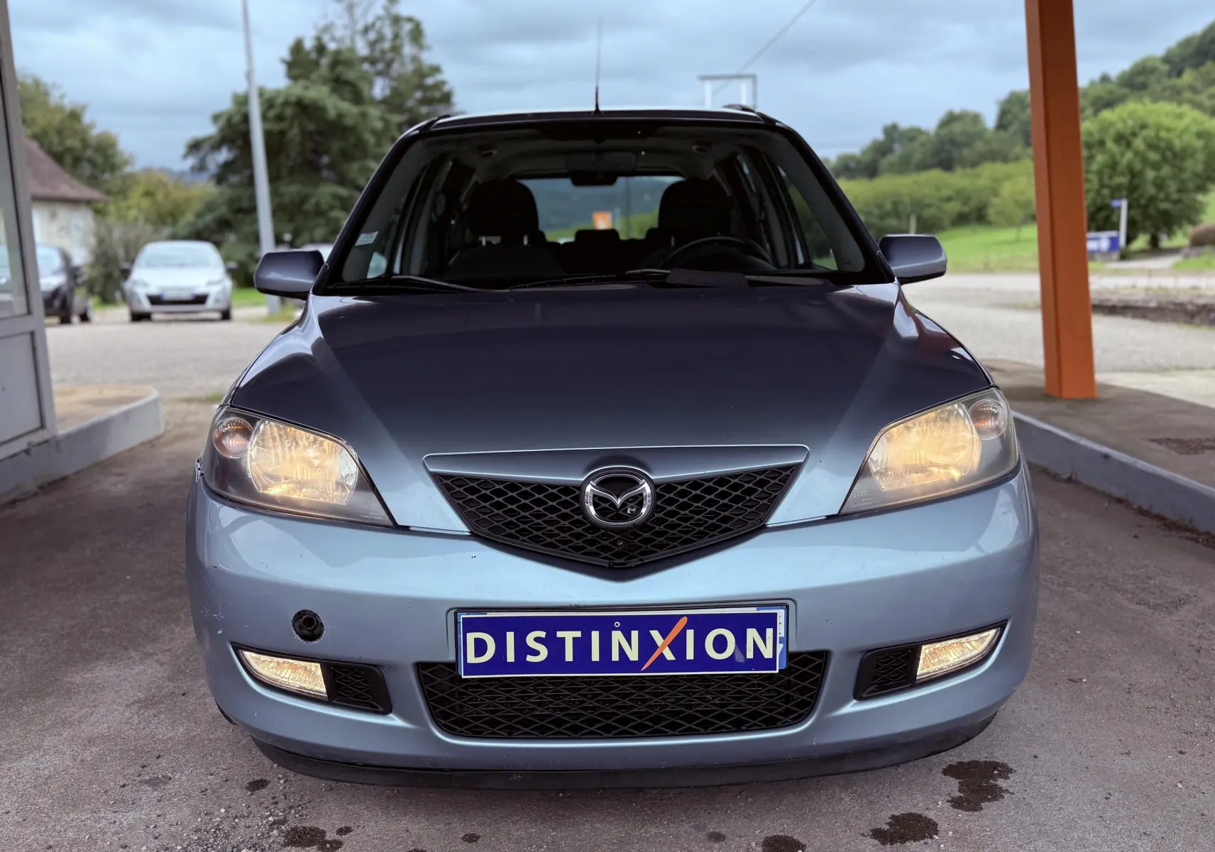 Vue frontale d'une Mazda2 gris clair avec phares allumés et plaque d'immatriculation Distinxion visible.