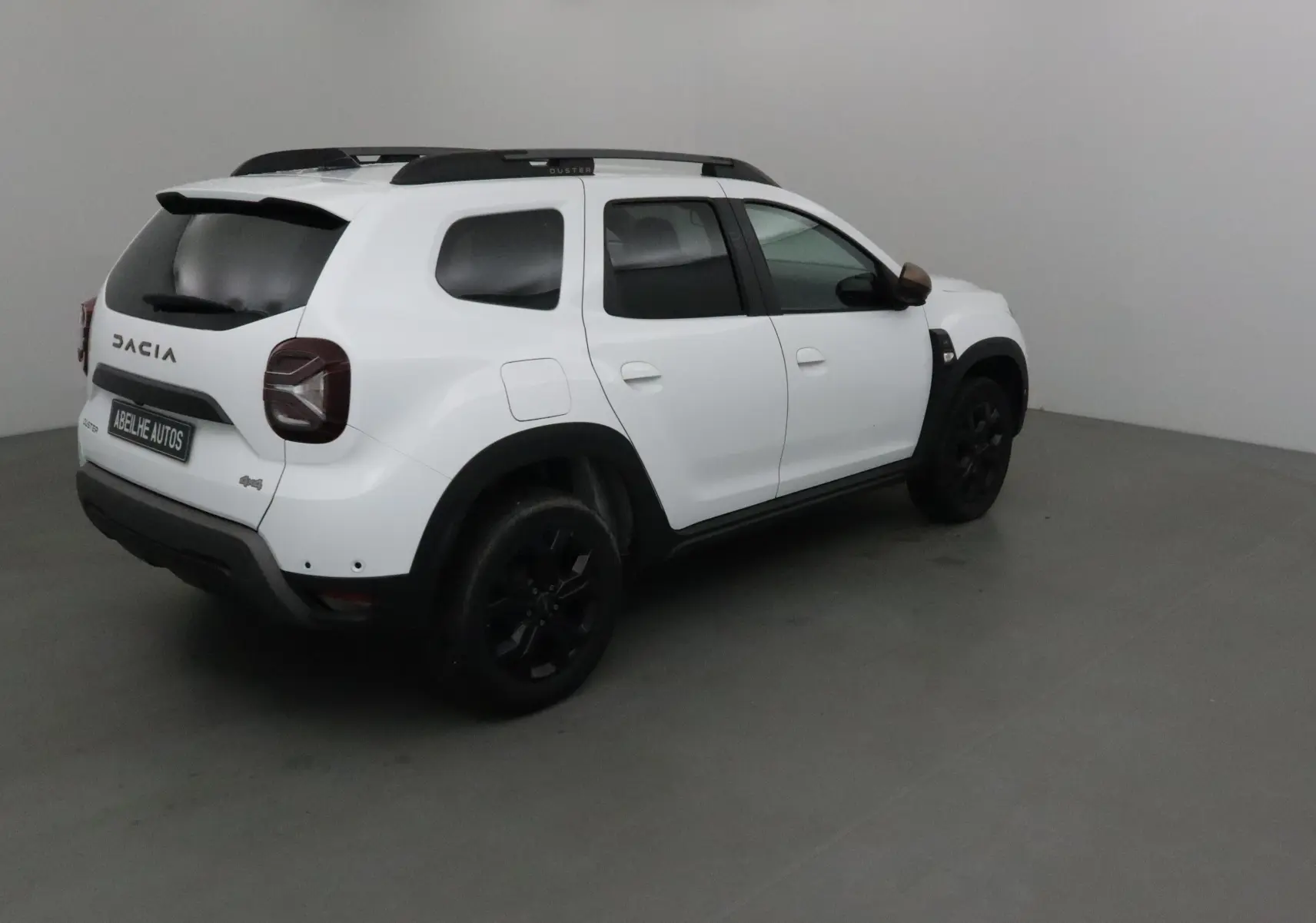 Vue 3/4 arrière droite d'un Dacia Duster 4x4 blanc 2024 avec jantes noires et barres de toit noires.