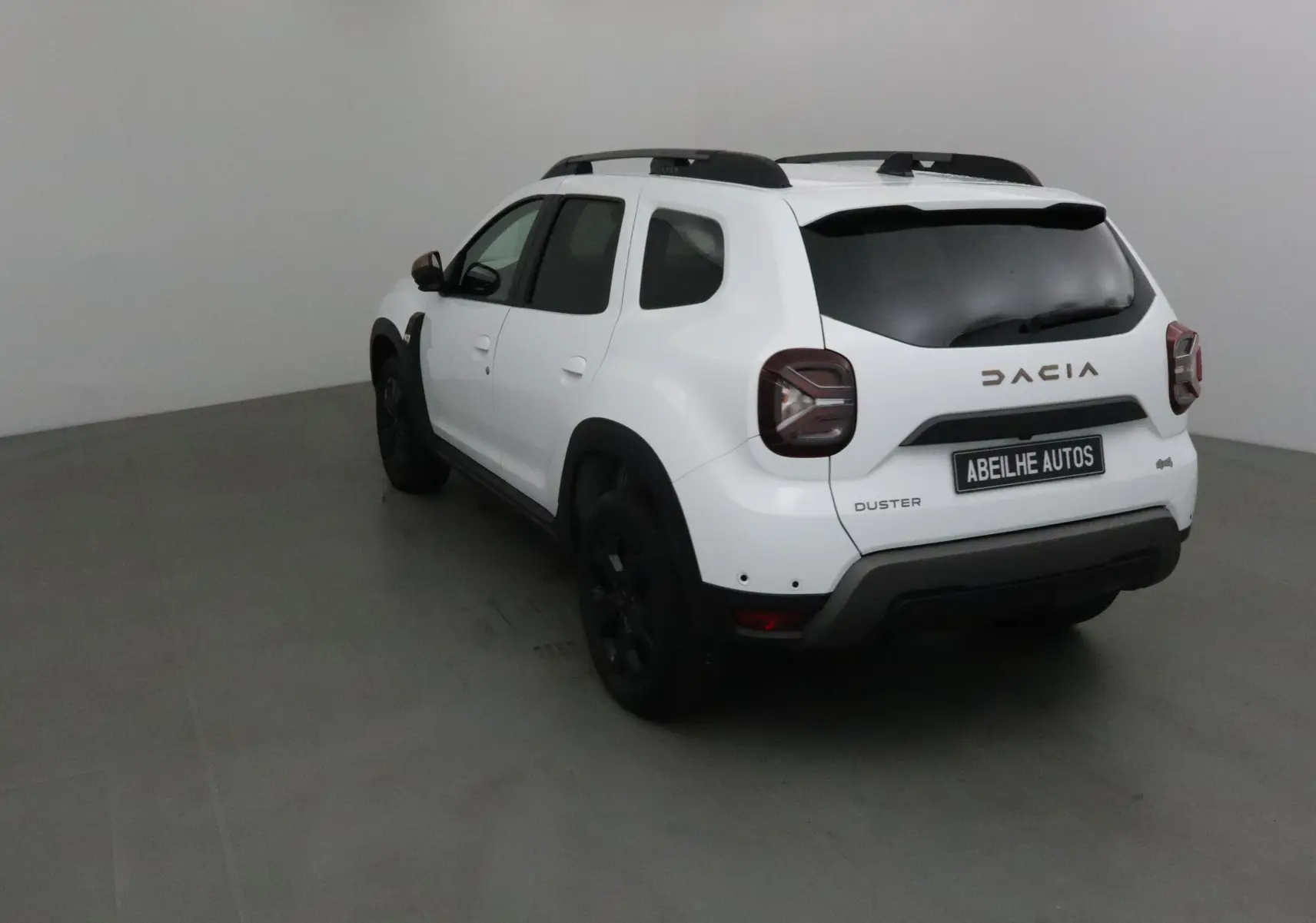 Vue 3/4 arrière droite d'un Dacia Duster blanc 2024 avec jantes noires et barres de toit noires.