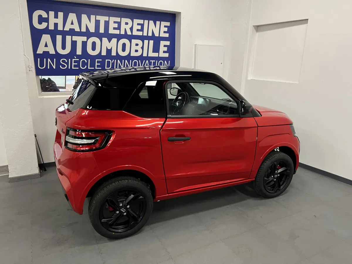 Vue côté droit arrière de la LIGIER JS50 BEV 8KWH ELITE rouge avec toit noir et jantes alu noires en intérieur showroom.