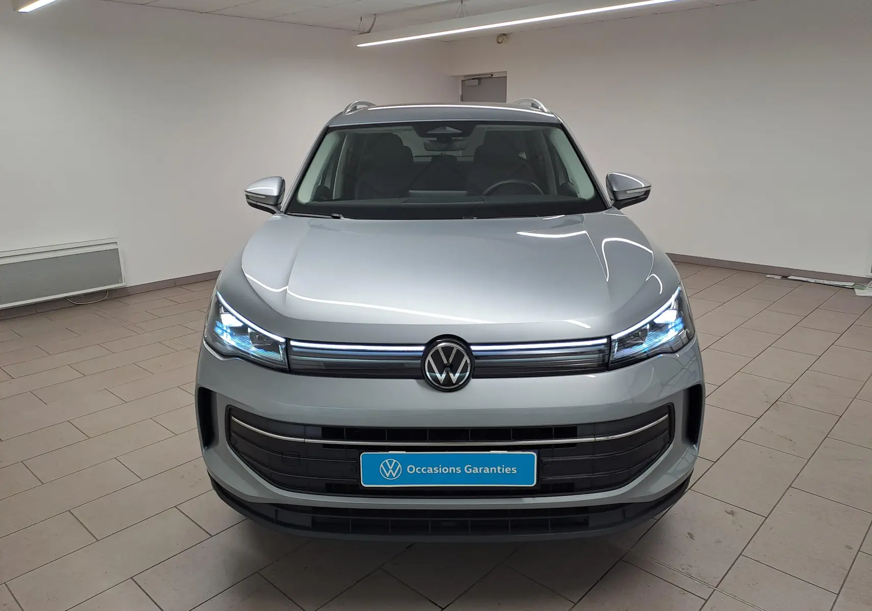 Vue frontale du Volkswagen Tiguan 2.0 TDI 150 DSG7 VW Edition en argent Dolomite avec phares LED allumés dans un showroom.