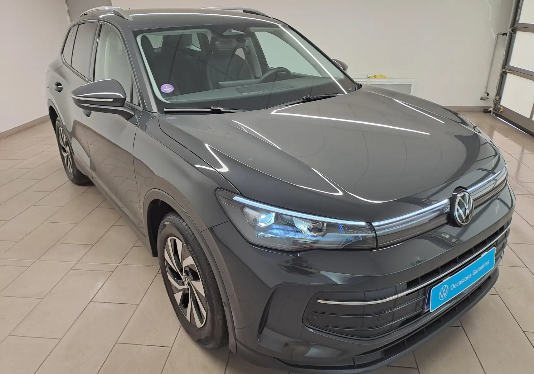 Volkswagen Tiguan 2024 gris Urano en 3/4 avant droit, avec phares allumés et crochet d'attelage escamotable visible.