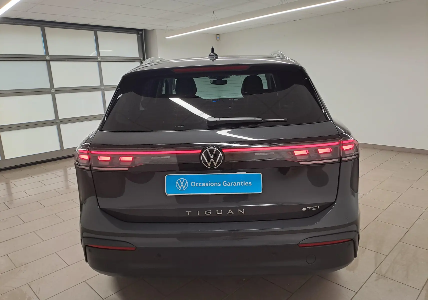 Vue arrière d'un Volkswagen Tiguan gris Urano 2024 avec feux allumés et plaque "Occasions Garantis" en intérieur showroom.