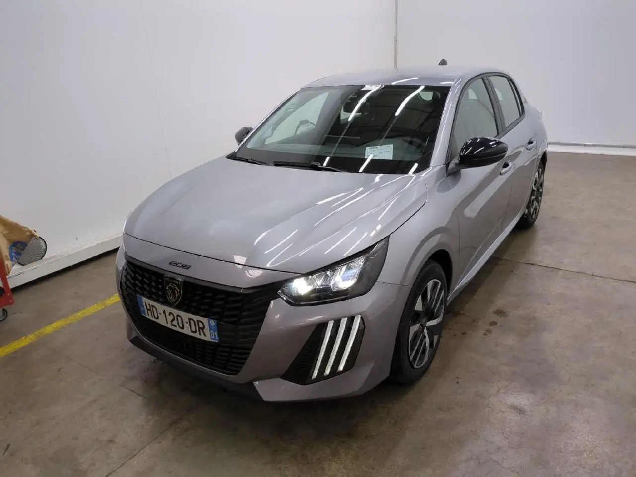 Peugeot 208 gris clair vue 3/4 avant droit avec calandre noire et feux LED allumés dans un garage.