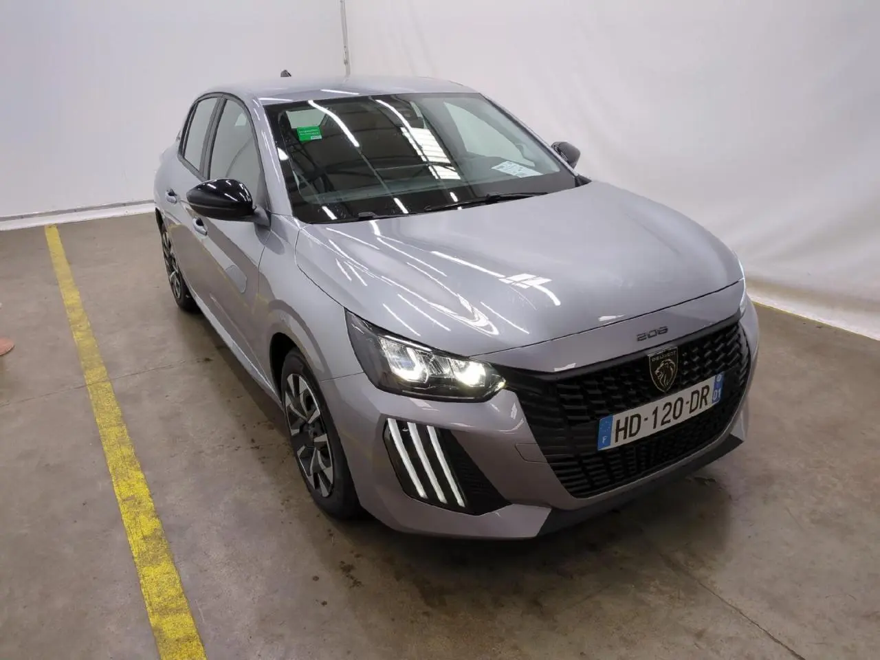 Peugeot 208 gris clair vue 3/4 avant droit avec calandre noire et feux LED allumés dans un garage.