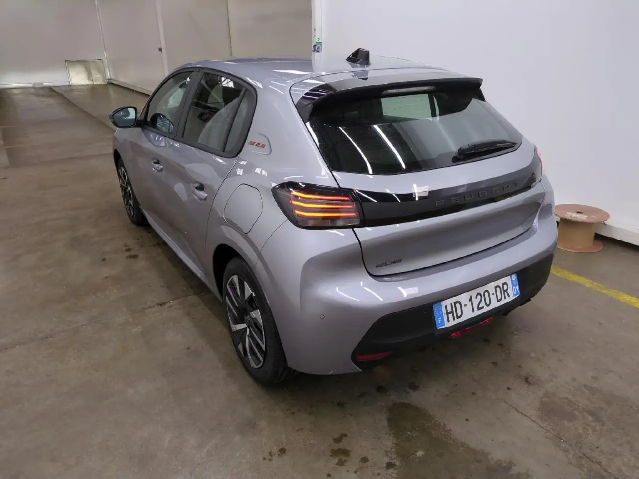 Vue 3/4 arrière droite d'une Peugeot 208 gris clair avec bandeau noir brillant entre feux et badge Style sur custode.