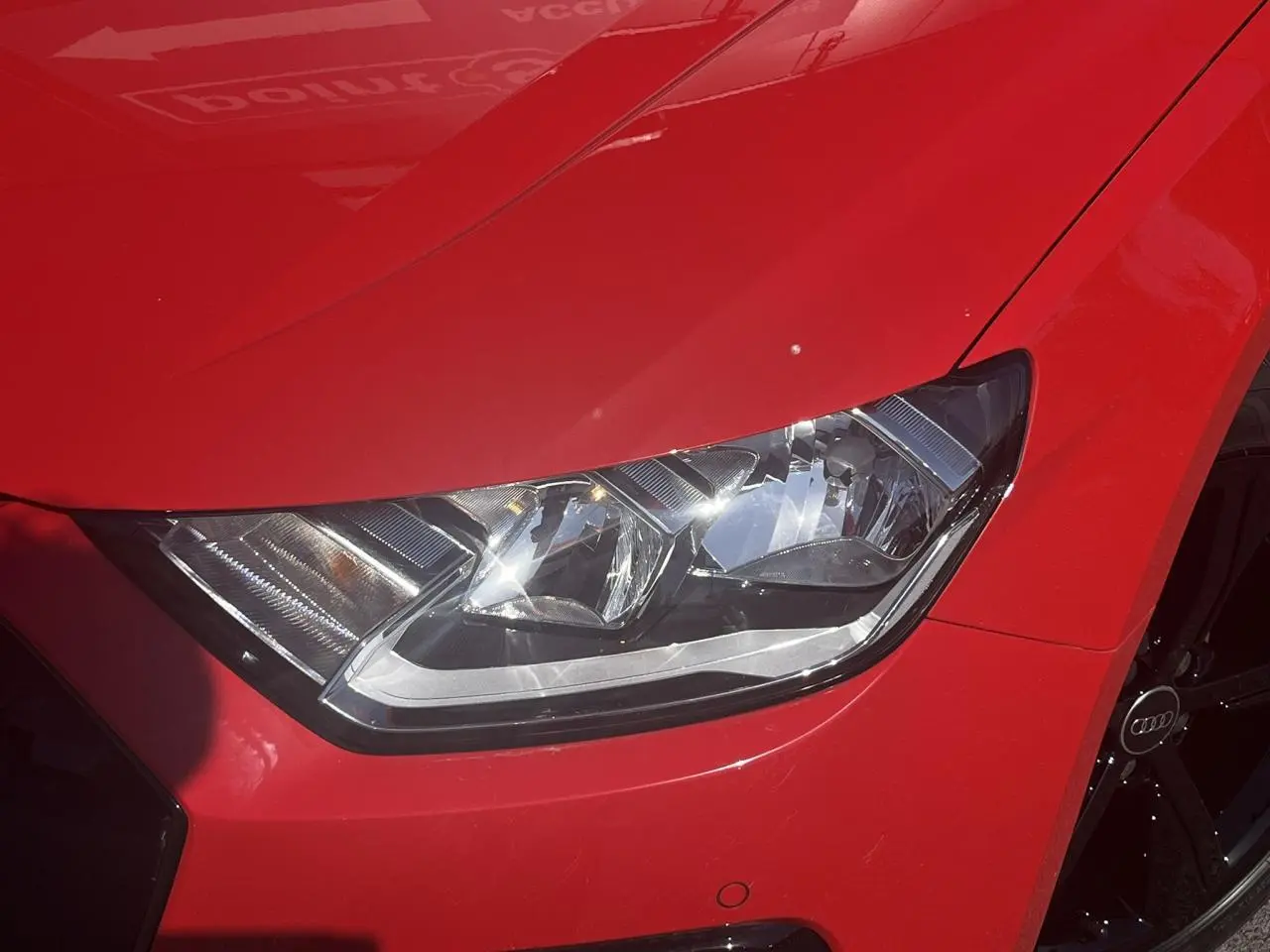Gros plan sur le phare avant droit et la jante noire d'une Audi A1 Sportback rouge Misano, version 25 TFSI 95 Advanced.