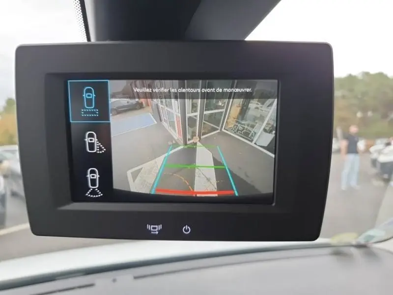 Écran de caméra de recul du Peugeot Partner blanc montrant l'arrière avec lignes de guidage et parking devant un bâtiment.