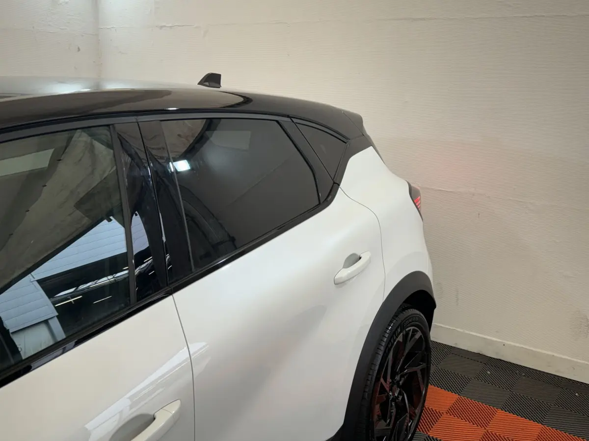 Vue côté gauche arrière du Renault Captur blanc avec toit noir et jantes noires sportives dans un intérieur lumineux.