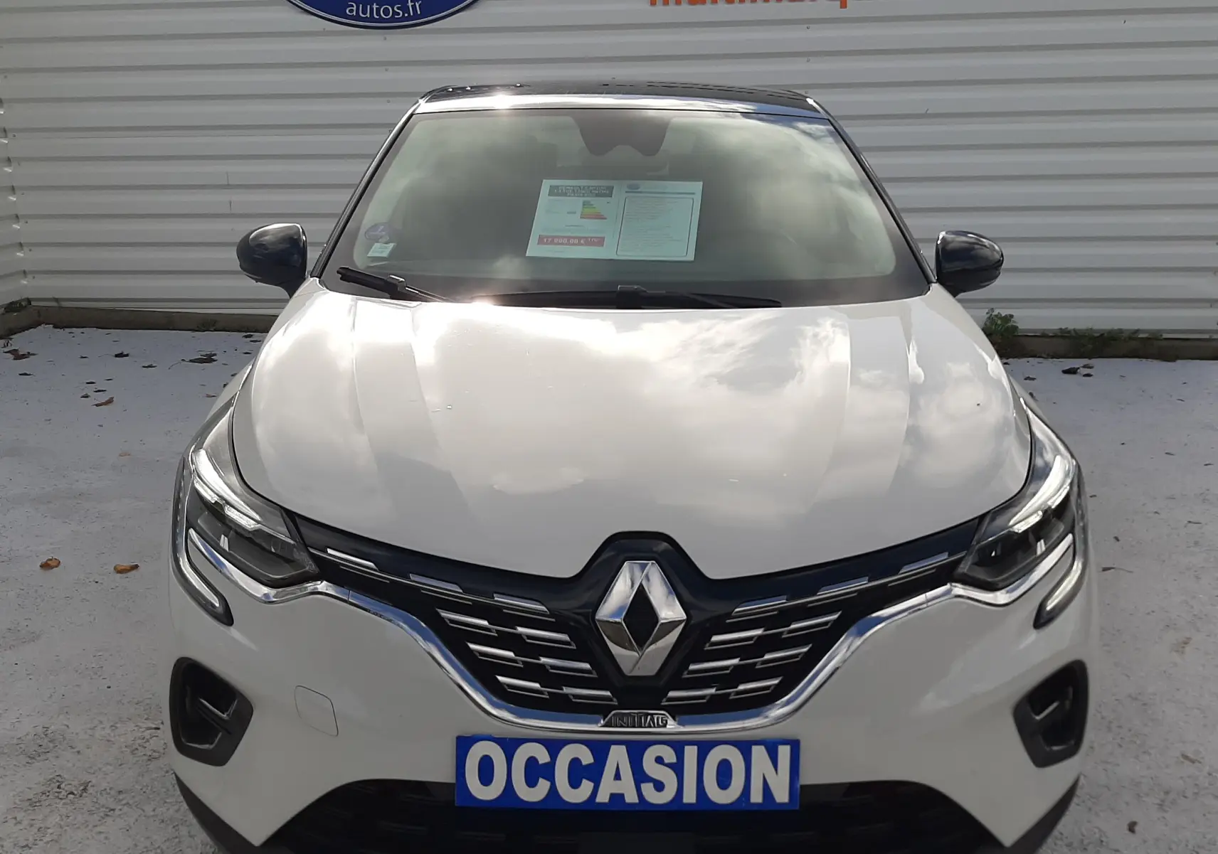 Vue frontale d'un Renault Captur blanc avec toit noir, affichant une plaque "OCCASION" devant un mur publicitaire.