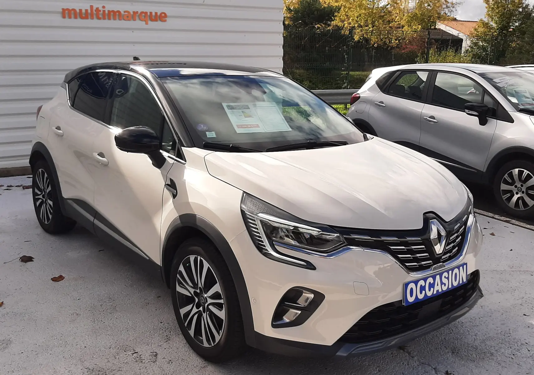 Renault Captur blanc avec toit noir en 3/4 avant droit, stationné devant un mur blanc avec panneau multimarque.