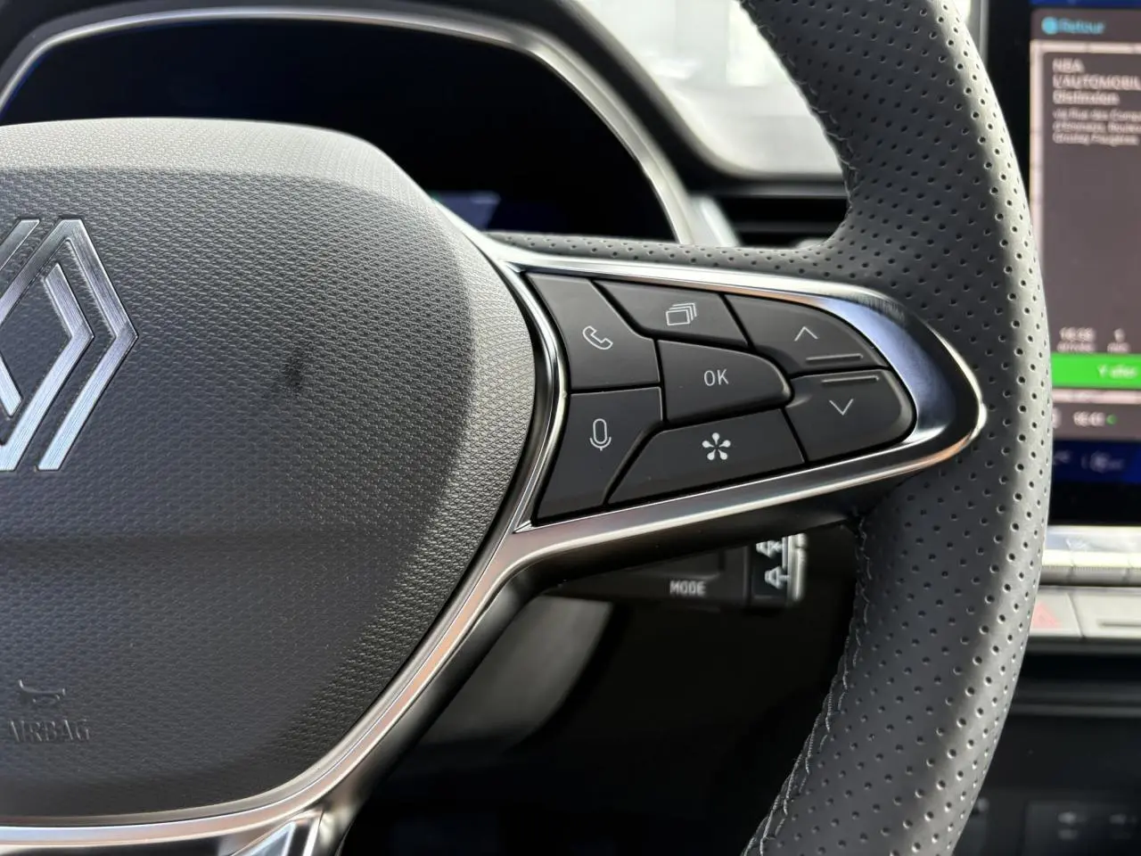 Gros plan sur le volant en cuir perforé de la Renault SYMBIOZ gris Rafale, avec commandes multifonctions et écran tactile en arrière-plan.