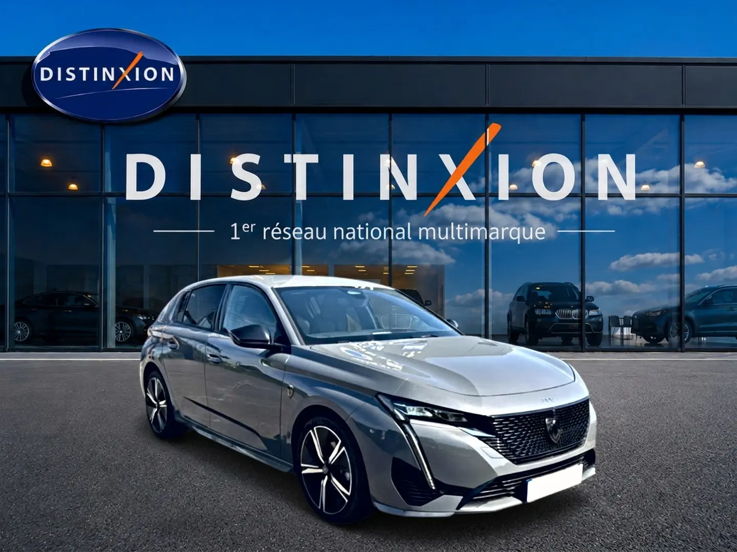 Peugeot 308 gris en 3/4 avant droit, avec calandre noire et jantes alliage distinctives, devant un showroom Distinxion.