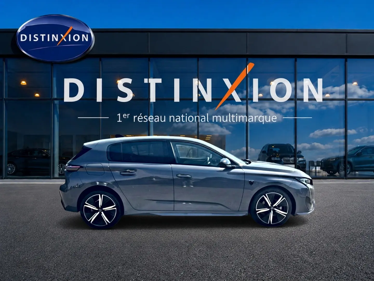 Profil droit d'une Peugeot 308 gris métallisé 2025 garée devant une vitrine avec logo Distinxion.